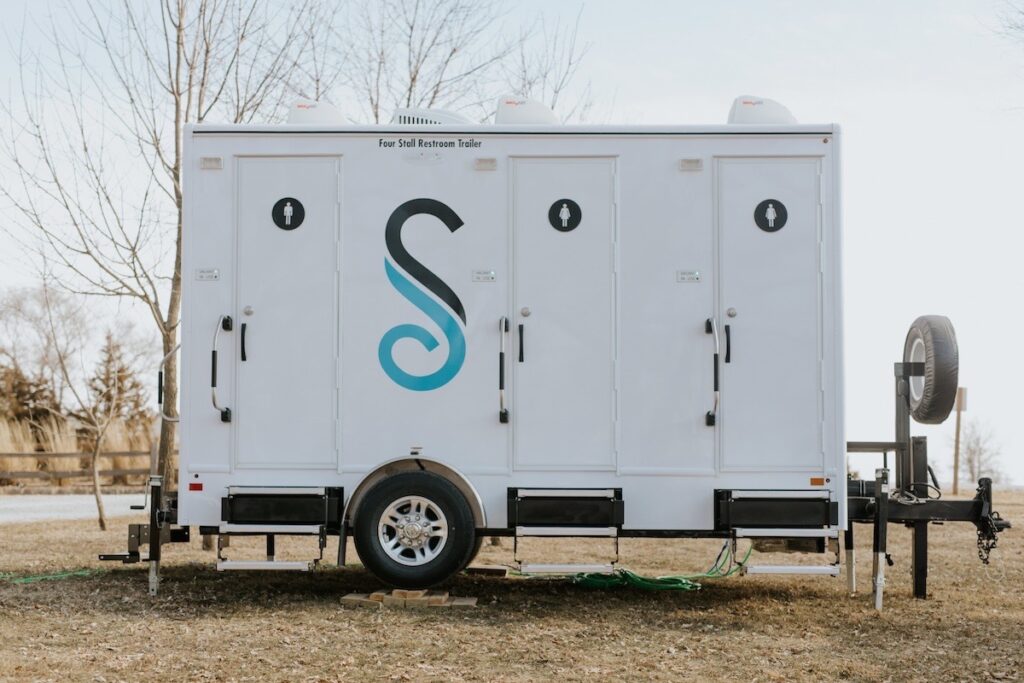 A white portable restroom trailer with four doors, each marked with a gender neutral restroom sign, is parked outdoors on a grassy area—offering essential comforts and rustic charm for any modern farm wedding.
