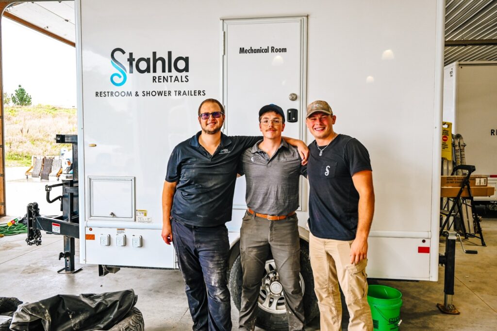 Three men stand arm in arm smiling in front of a Stahla Rentals restroom and shower trailer inside a covered area.