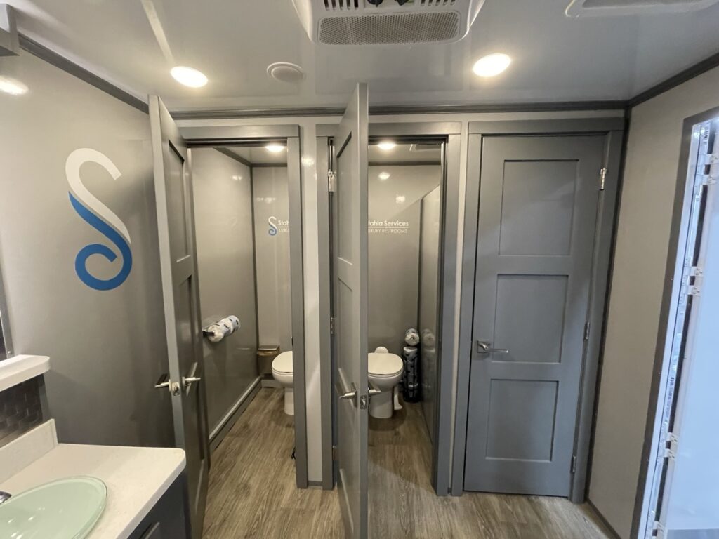 A clean, modern restroom with two stalls featuring toilets, a sink on the left, and gray doors and walls—perfect for adding essential comforts to your modern farm wedding or event with subtle rustic charm.