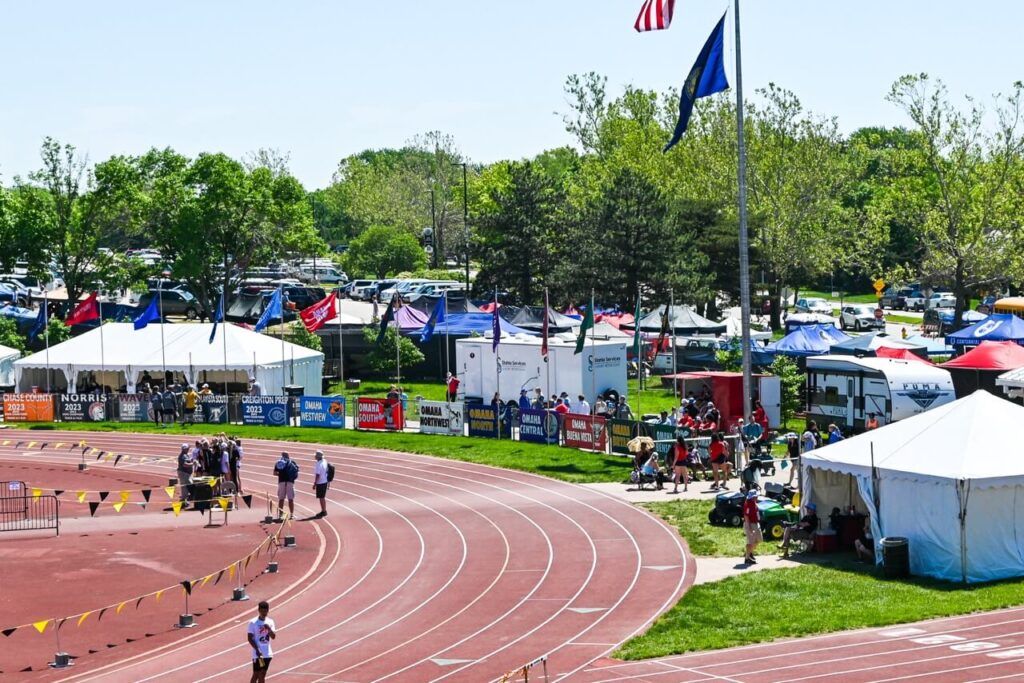 an outdoor track meet at the nsaa state track event features runners on the curved track, spectators, tents, and flags alongside. trees and parked vehicles are visible in the background under a clear sky, enhancing the attendee experience with convenient restroom trailers on site.