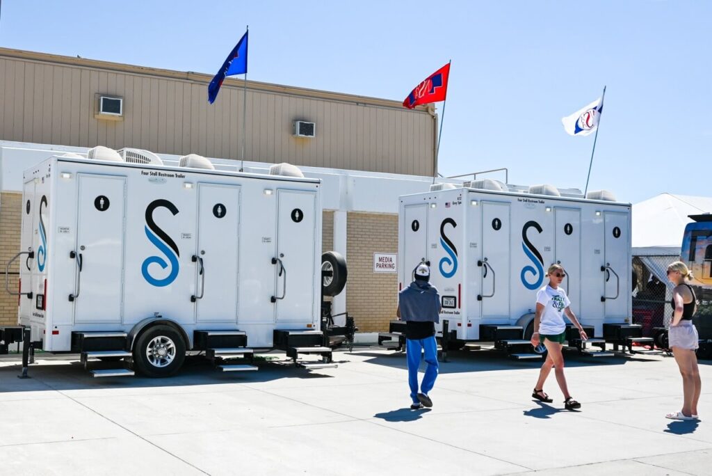 attendees walk past a row of portable restroom trailers with blue and white graphics, set up outside a large building under a clear sky with flags on top, enhancing the attendee experience at the nsaa state track event.