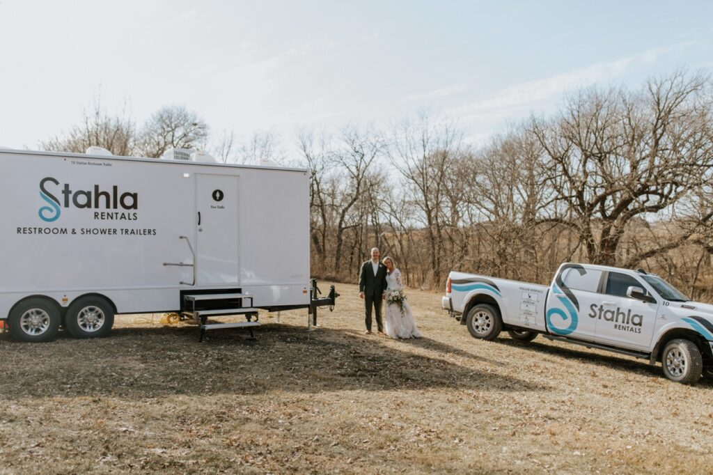 A bride and groom stand outside next to a stahla rentals restroom and shower trailer, enhancing the guest experience, alongside a branded pickup truck on a grassy, wooded lot.