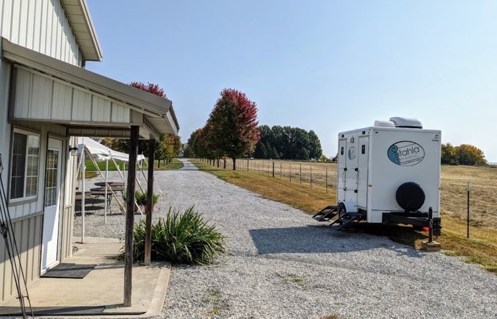 Portable restroom trailer by countryside building with trees.