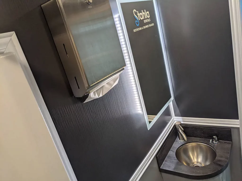 a modern, compact 3 stall restroom trailer rental with a metallic sink, dark textured walls, a mirror with an illuminated frame, and small cabinets labeled "stahl" above.