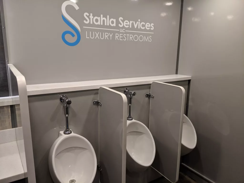 three white urinals with privacy dividers in a well maintained restroom trailer displaying the logo "stahla services llc luxury restrooms" on the wall.