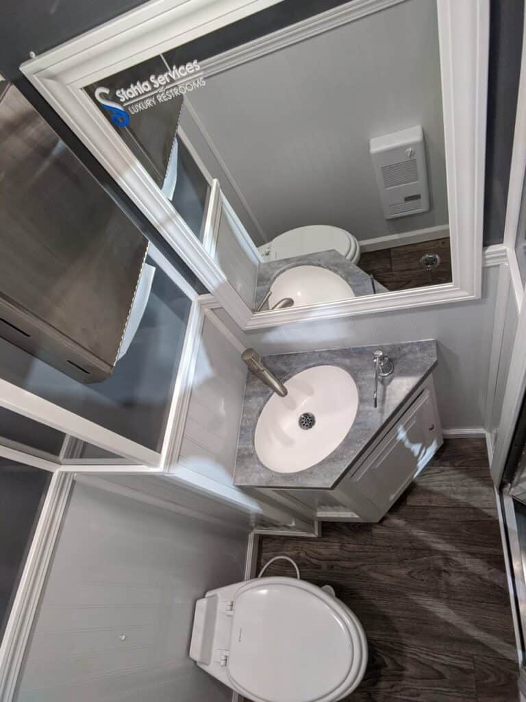 a small modern 2 stall restroom trailer rental featuring a white toilet, a sink with a cabinet, wood flooring, and mirrored walls, viewed from above.