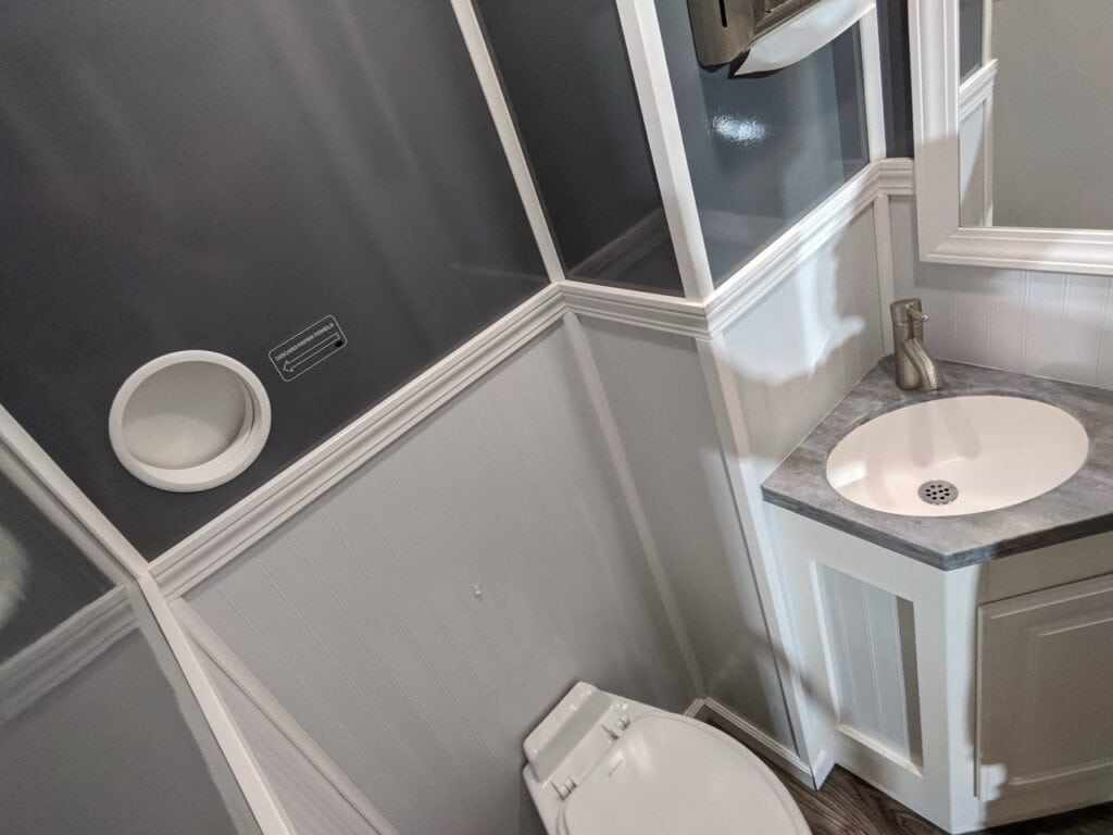 interior of a 2 stall restroom trailer with dark gray walls, white paneling, rectangular vanities with sinks, and toilets next to wall mounted round mirrors.