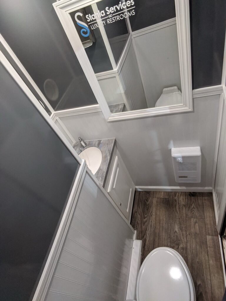 interior of a small, modern bathroom with gray walls, wooden floor, a white sink, and a toilet, viewed from above. a sign reads "2 stall restroom trailer rental luxury restrooms.
