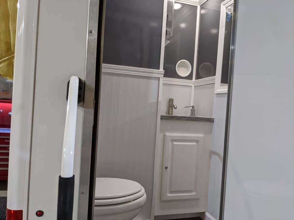 interior of a compact 2 stall restroom trailer rental featuring a white toilet, small sink with a mirror above it, and a shower stall visible through a glass door.