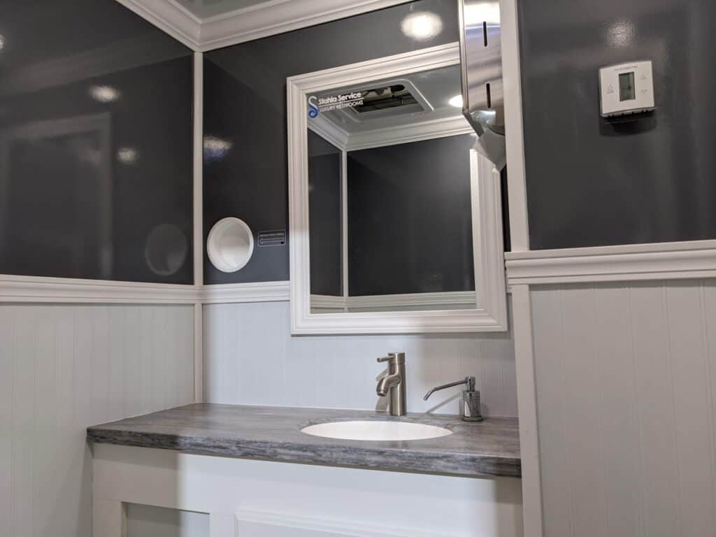 a modern bathroom vanity featuring a white sink, chrome faucet, and a mirrored medicine cabinet with a small shelf and multiple switches on the gray walls of a 2 stall restroom trailer rental.