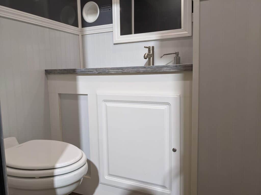 a small 2 stall restroom trailer with white paneling, featuring a toilet next to a cabinet with a built in sink and faucet under a window.