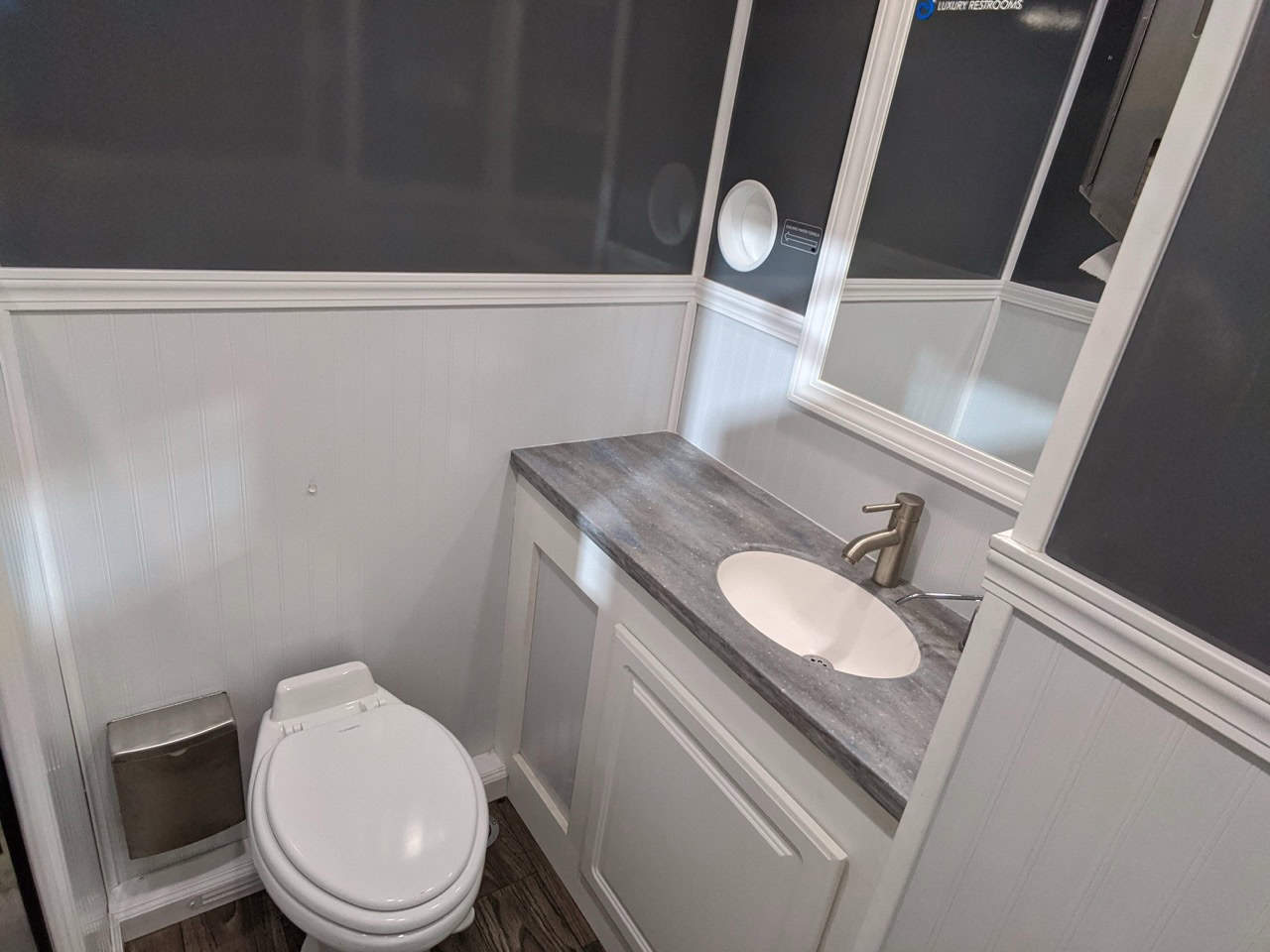 a small modern 2 stall restroom trailer rental with white walls, a gray vanity with an integrated sink, a toilet, and a mirrored medicine cabinet.