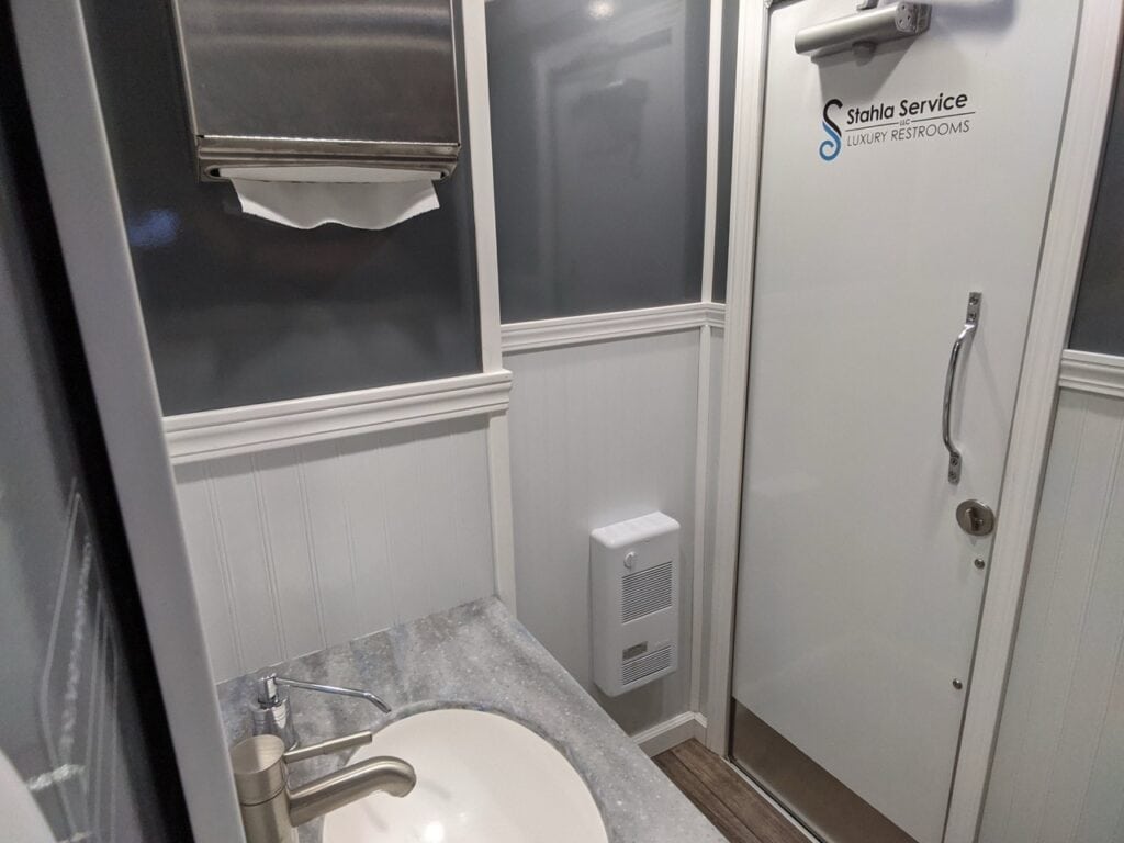 interior of a modern luxury 2 stall restroom trailer rental featuring a sink, gray countertop, cabinet, and a towel dispenser, with a door labeled "stadia service.