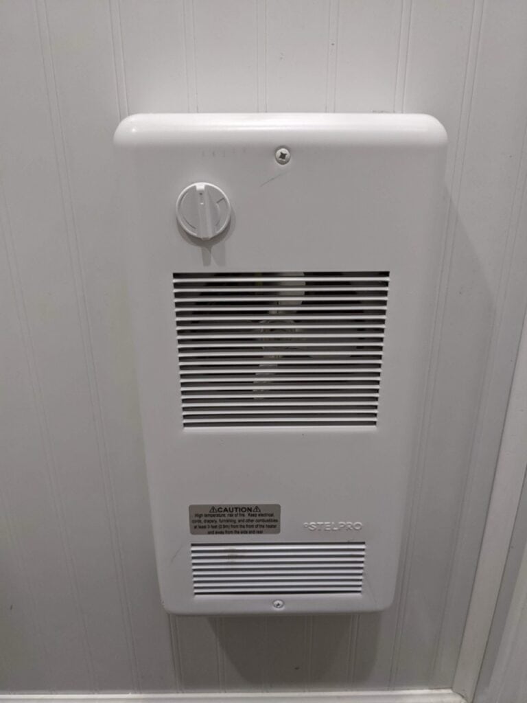 wall mounted white bathroom fan heater with dial, vent slats, and a caution label, installed on a white paneled wall in a 2 stall restroom trailer rental.