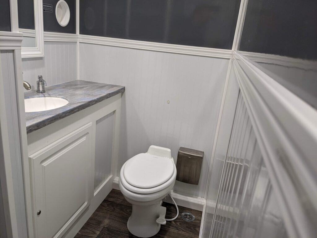 a small 2 stall restroom trailer rental featuring a white toilet beside a sink cabinet with a marble countertop, and white paneled walls.