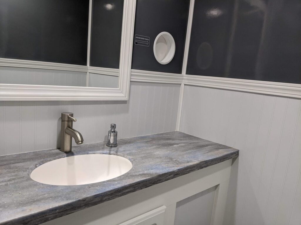 a modern 2 stall restroom trailer rental with a white sink, gray marble countertop, white wooden paneling, and a round mirror against a dark blue wall.