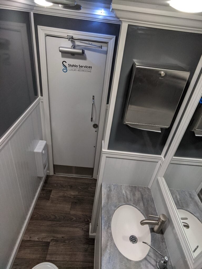 interior of a modern bathroom with white walls, a wooden floor, a white sink, and a stainless steel mirror, featuring a door labeled 'stahla services 2 stall restroom trailer rental'.