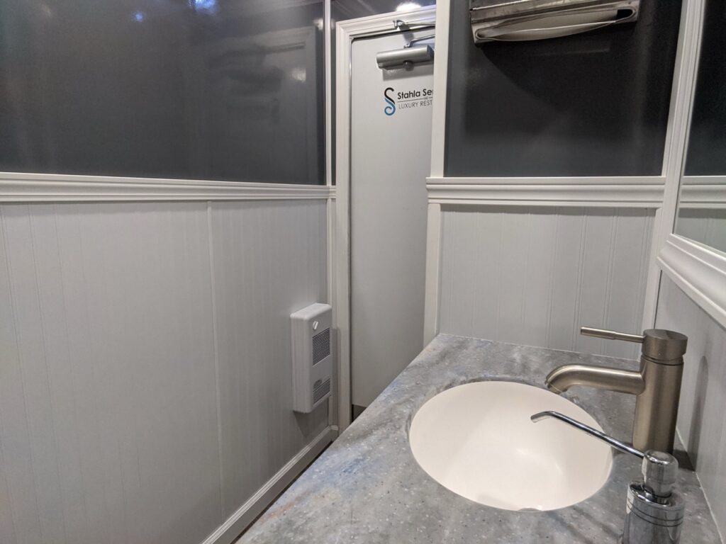 a small 2 stall restroom trailer rental corner with a white sink, silver faucet, and grey countertop, featuring a white paneled wall and a sliding shower door.