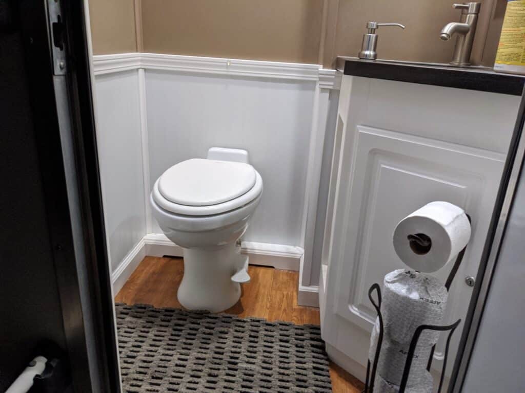 a small bathroom with a white toilet, a white cabinet under a sink, and a metal holder with a roll of toilet paper near the toilet, reminiscent of what you'd find in a well maintained stall restroom trailer rental.