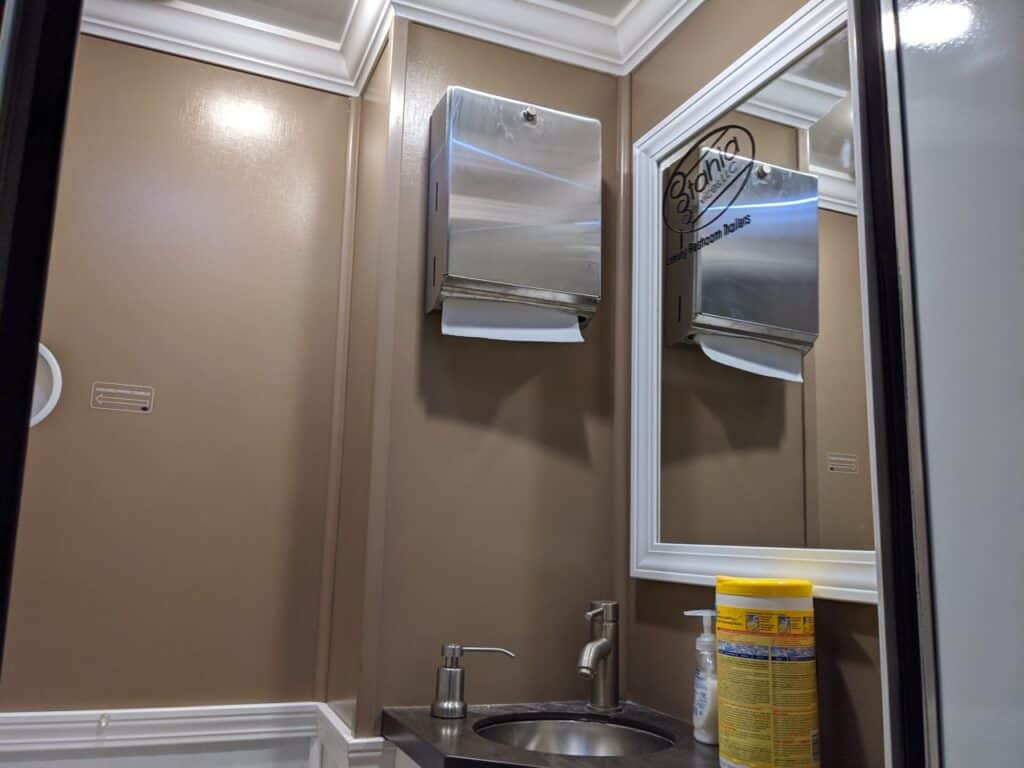 a 1 stall restroom trailer rental features a bathroom with beige walls, a paper towel dispenser, a mirror, a sink with a soap dispenser, and a container of disinfecting wipes on the countertop.