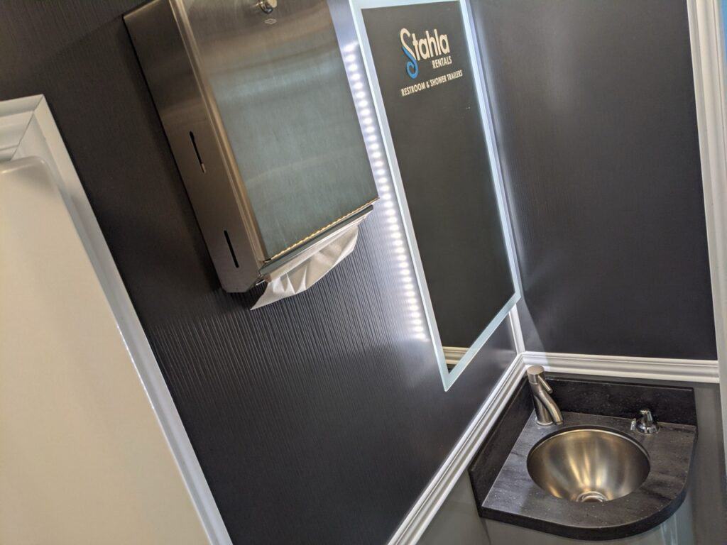 a small bathroom in the stall restroom trailer rental features a rectangular mirror, a stainless steel sink, and a wall mounted paper towel dispenser. the mirror has some text at the top reflecting the restroom service branding.