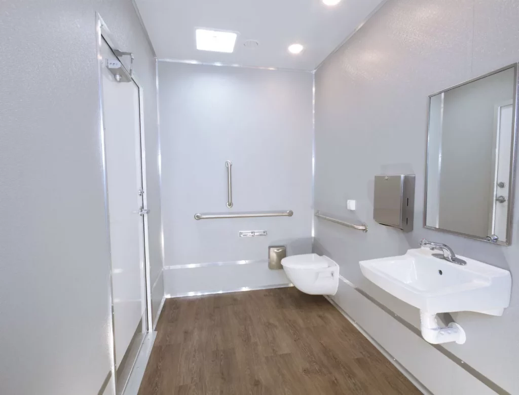 a modern, accessible restroom trailer interior featuring a toilet with grab bars, a sink, mirror, and a paper towel dispenser, with clean white walls and wooden flooring.