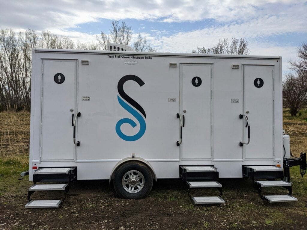a 3 stall shower trailer with three doors equipped with ramps, featuring male and female restroom signs and a large "s" logo in the center. this restroom trailer is situated outdoors near a forested area.