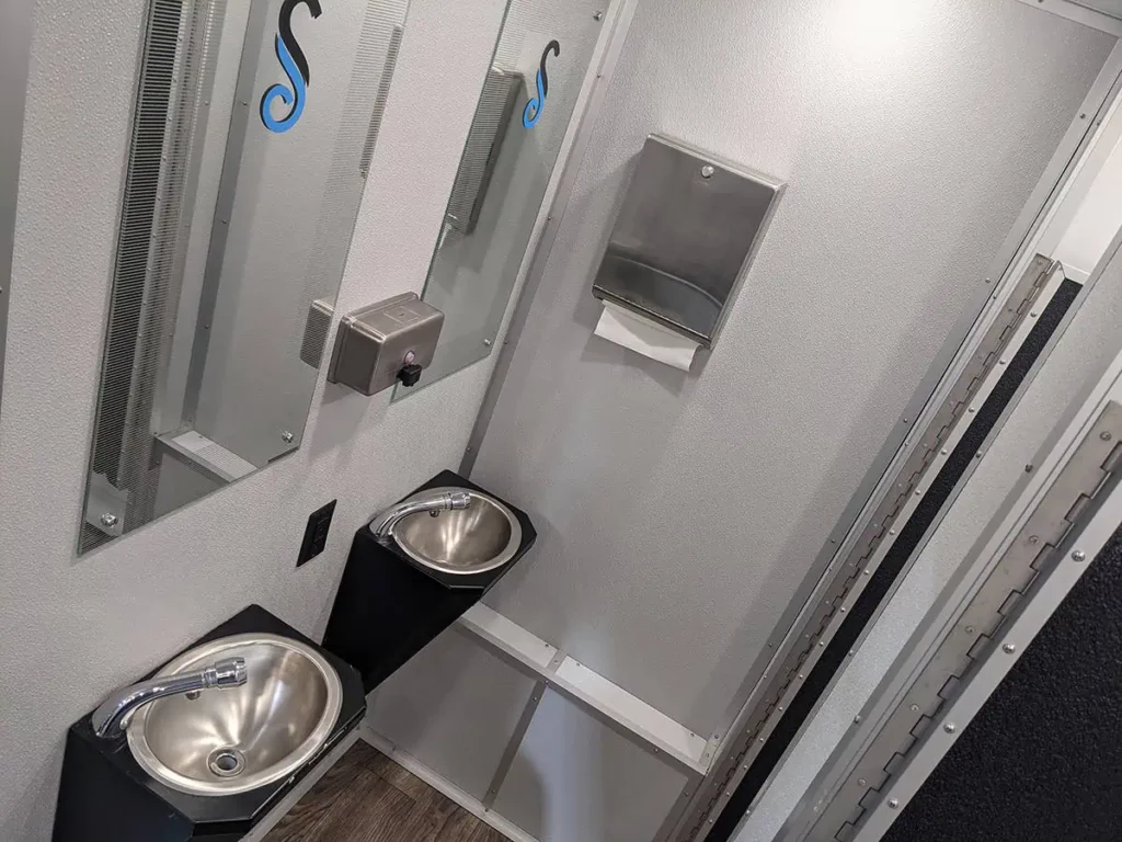 modern public restroom interior with two stainless steel sinks, soap dispensers, and wall mounted trash bins, featuring gray walls and a mirror in an 8 stall restroom trailer rental.