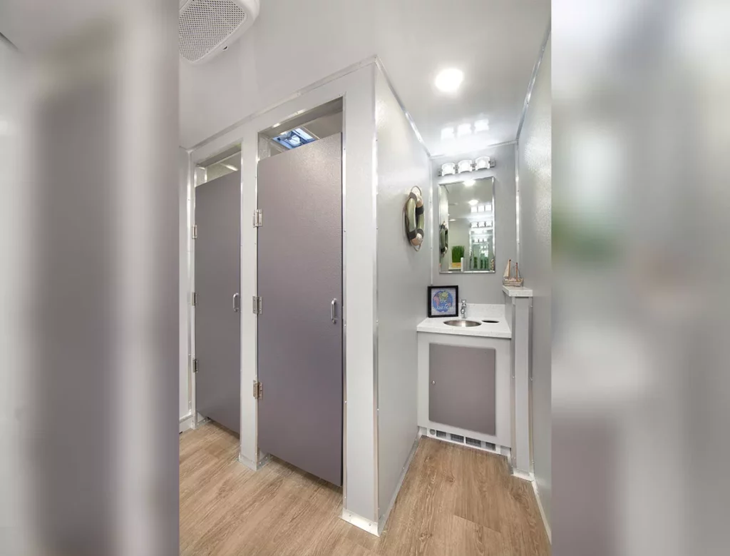 modern 10 stall restroom trailer interior with led lit mirrors and gray doors. a small vanity area with a digital display is visible. neutral, bright color scheme.
