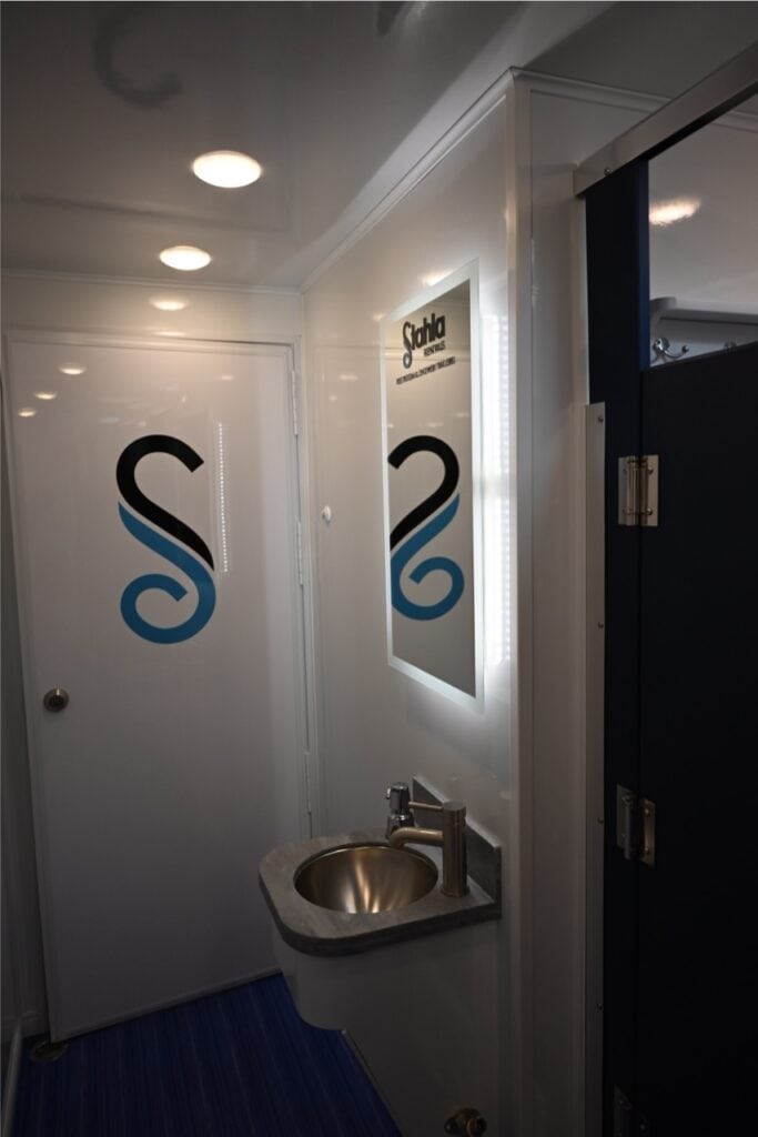 interior of a compact bathroom with white walls, featuring a round sink, mirror, and a reflective door marked with a stylized "s" logo on an 8 station shower trailer.