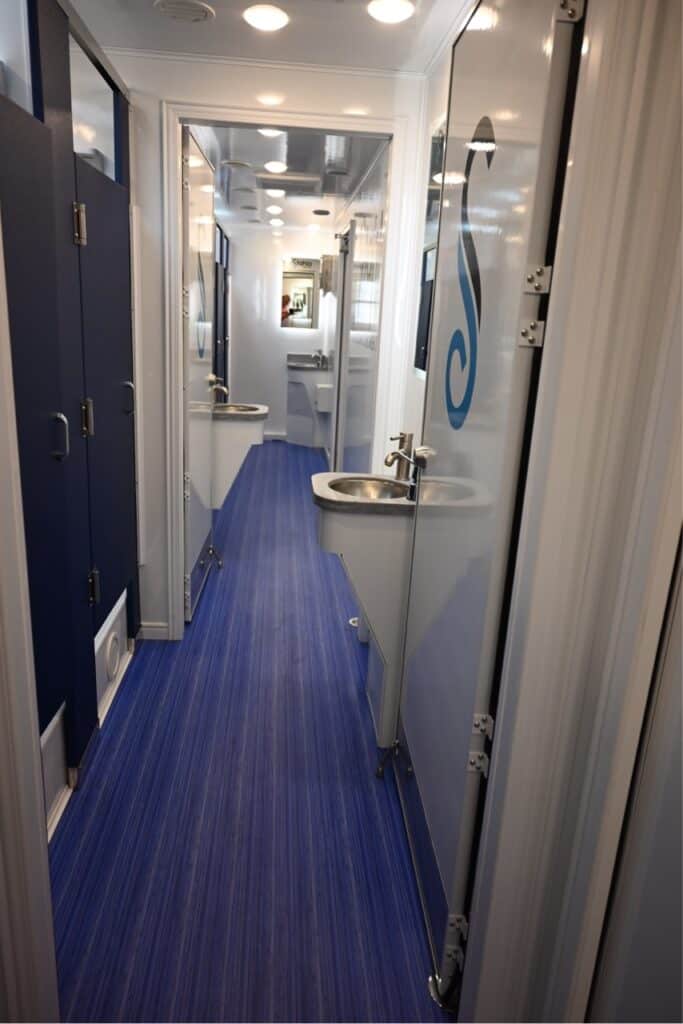 interior view of a narrow 8 station shower trailer with blue flooring, multiple doors, and a visible sink.
