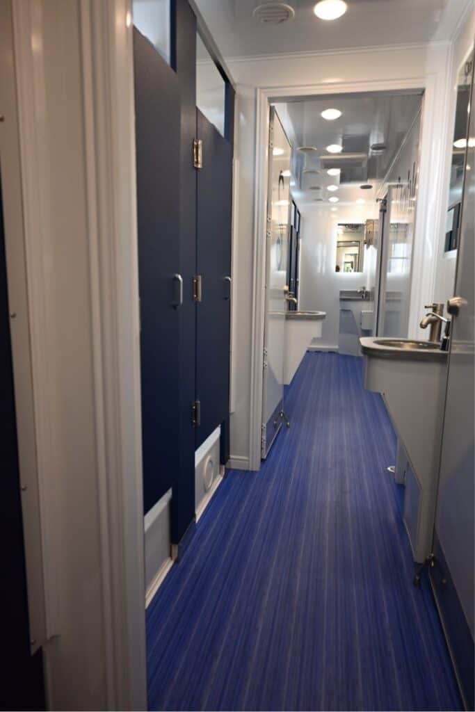 narrow train corridor with blue carpet and doors on one side, intersecting a perpendicular hallway with an 8 station shower visible.