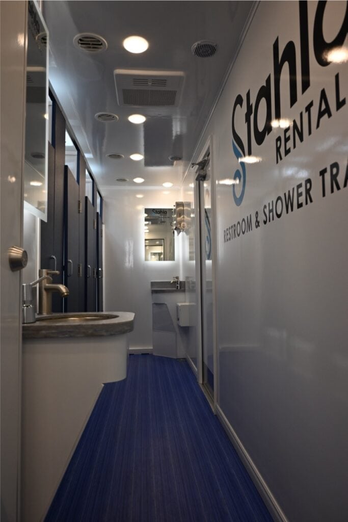 interior of a modern mobile shower trailer featuring a sink, mirrors, and entry to toilet stalls, with a blue floor and metallic walls.