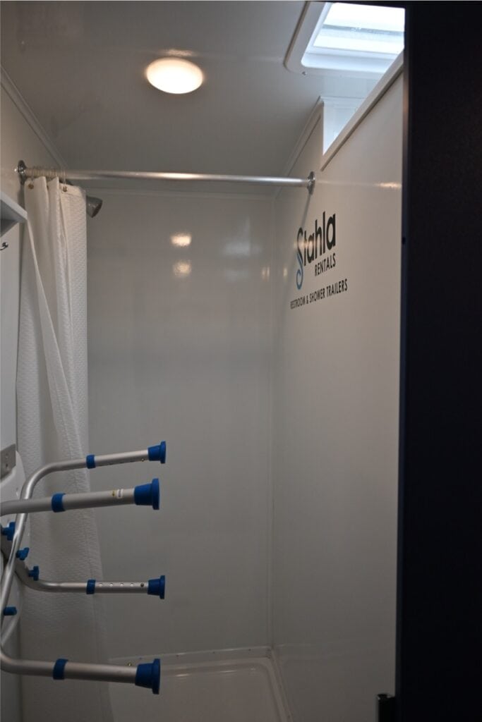 interior of a small, modern 8 station shower trailer with white walls and a blue grab bar, labeled "stahla rentals restroom shower trailers.