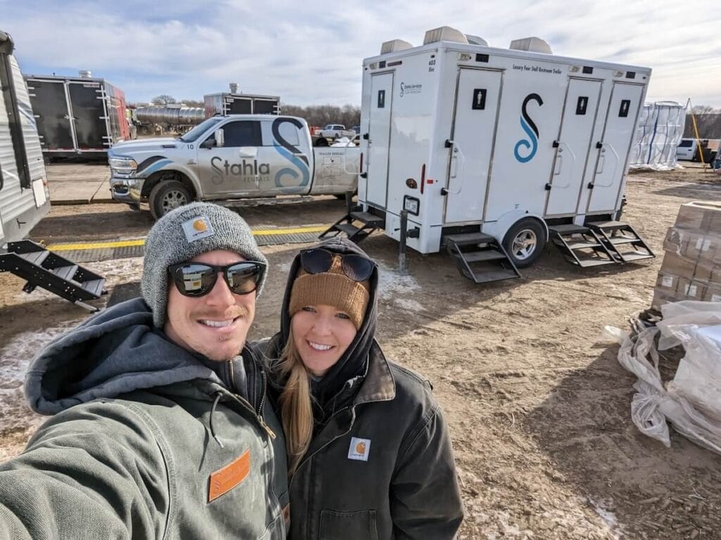 Couple selfie with portable restroom trailers in background.