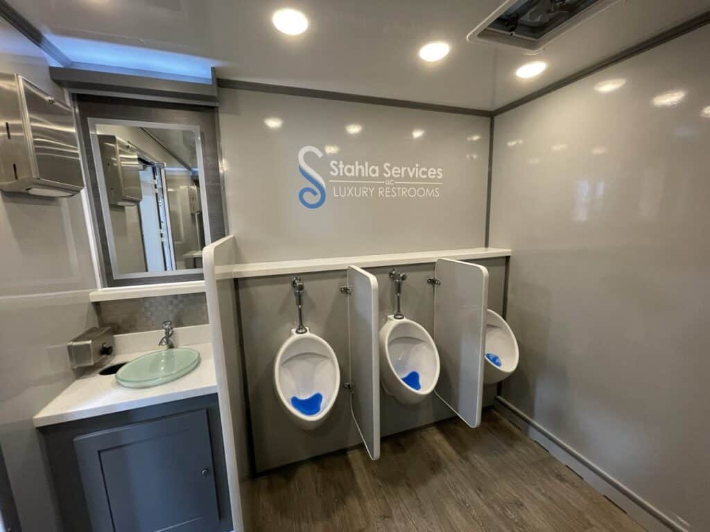 interior of a luxury 10 stall restroom trailer featuring three urinals and a sink, with "stahla services luxury restrooms" branding on the wall.
