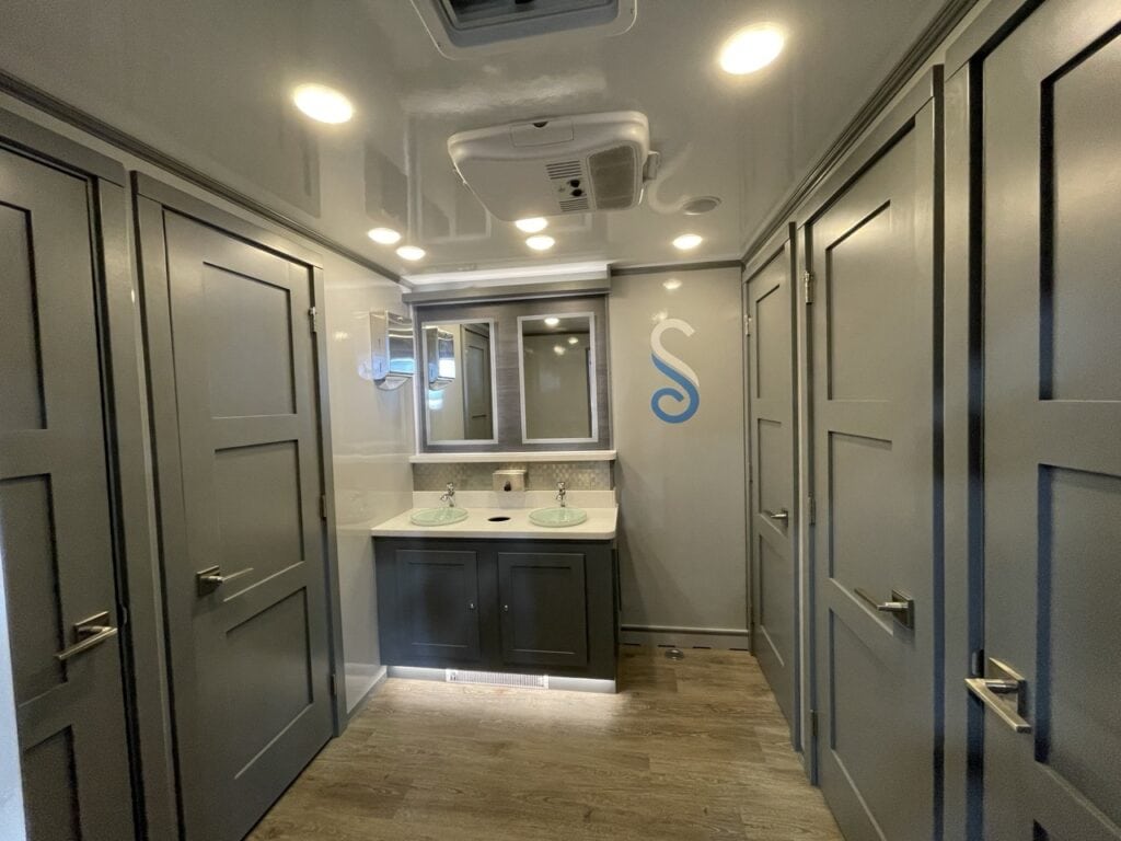 interior of a modern 10 stall restroom trailer featuring two sinks, gray cabinets, a large mirror, and wooden flooring.