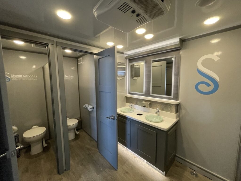 interior of a modern 10 stall restroom trailer with sinks, mirrors, and gray walls marked with a blue "s" logo.