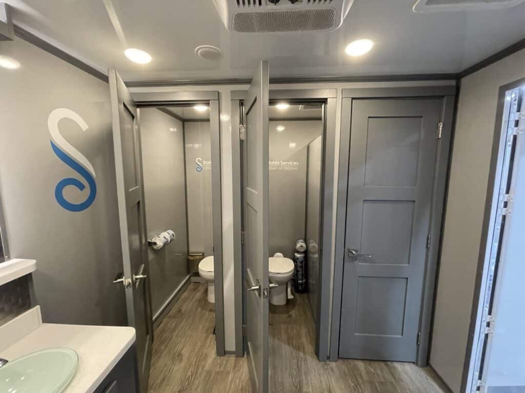 interior of a 10 stall restroom trailer with reflective sliding doors, a toilet, a sink with a mirror, and various toiletries.
