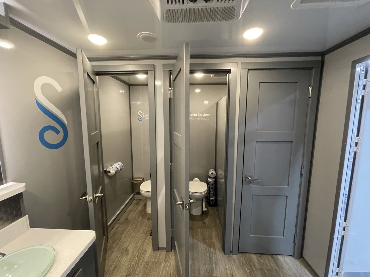 a clean, modern 6 stall restroom trailer featuring three stalls with a toilet, a urinal in another, and the third containing a sink area with a mirror. light grey walls and doors are prominent throughout.