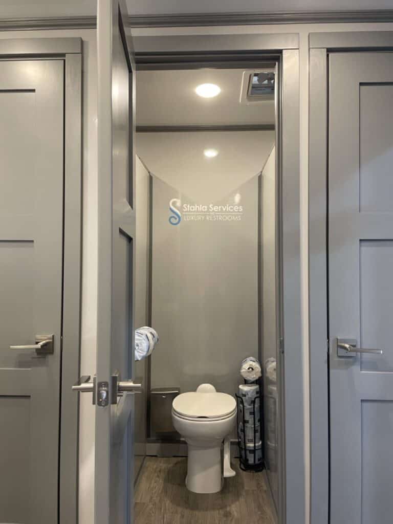 modern restroom interior with an open door, white toilet, and a stack of towels; signage reads "stahla services 10 stall restroom trailer" above the door.