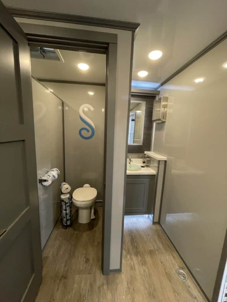interior of a 10 stall restroom trailer with frosted glass doors marked with an 's', featuring toilets, sinks, mirrors, and towel racks.