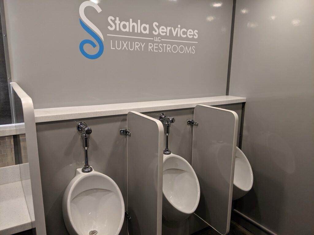 interior of a 10 stall restroom trailer showing three white urinals with dividers, and a logo of stahla services llc on the wall stating "luxury restrooms.