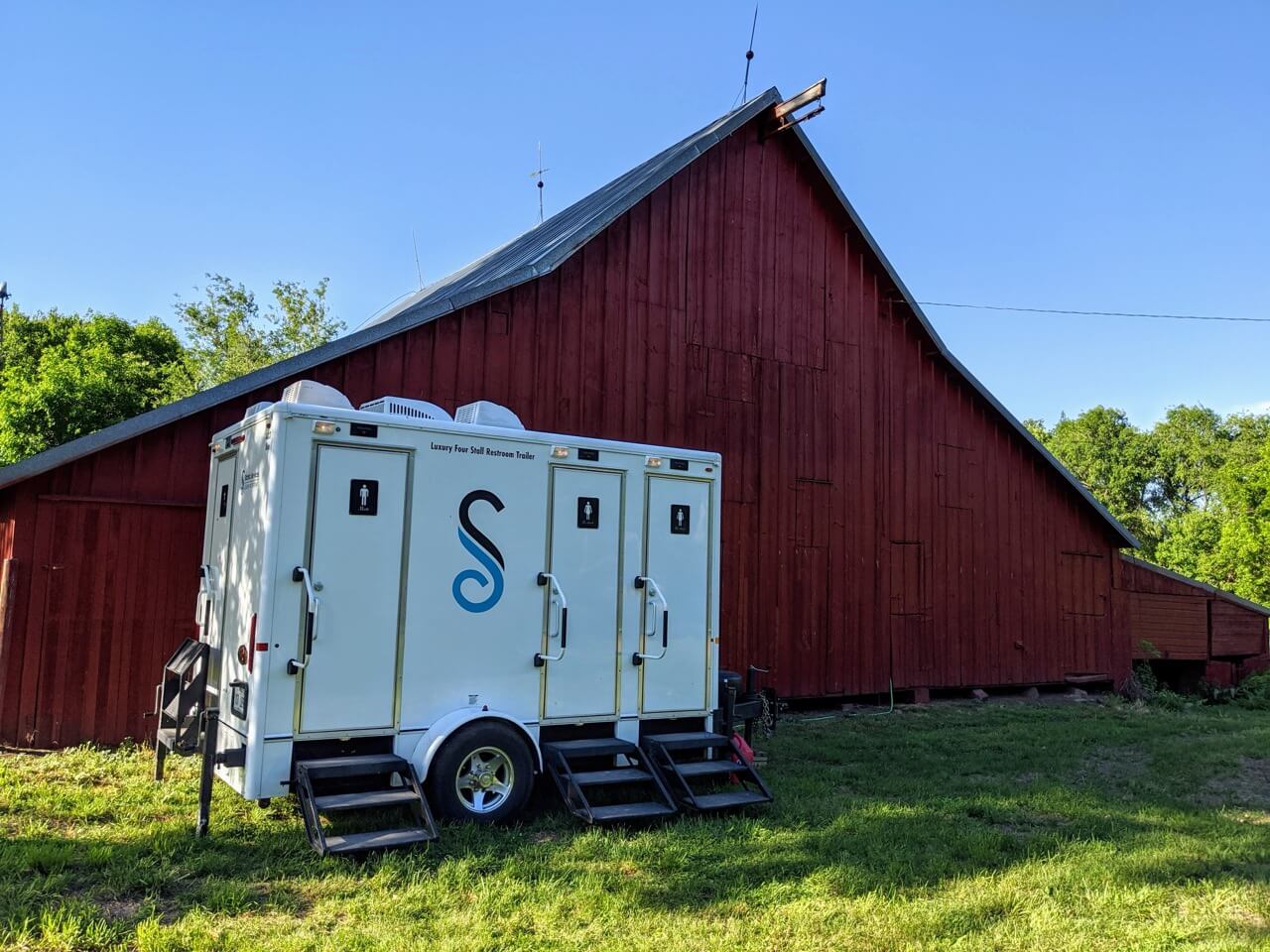a white 4 stall restroom trailer rental is parked on grass next to a large red barn under a clear blue sky.