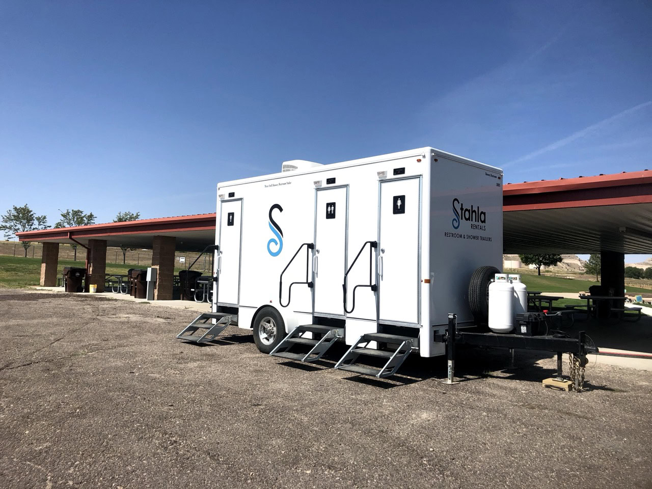 a portable 3 stall shower restroom trailer is parked on asphalt near a park shelter, equipped with steps, ventilation, and stahla rental branding, set against a clear sky backdrop.