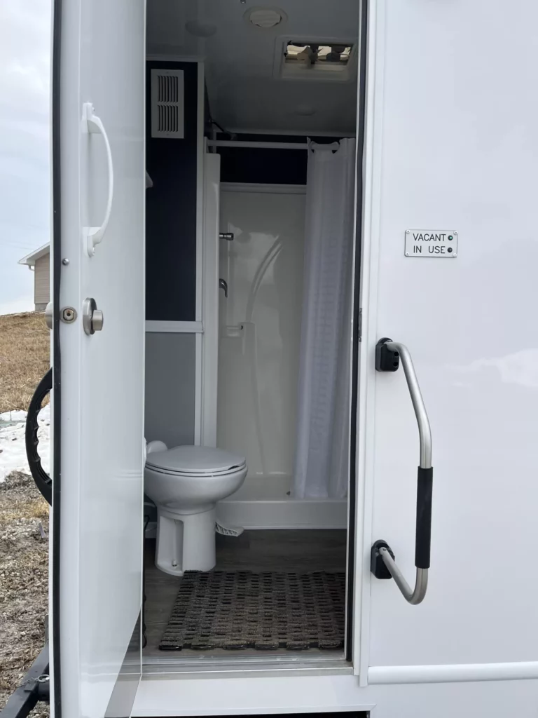 open door of a restroom trailer showing an interior with a toilet, shower, and occupancy indicator.