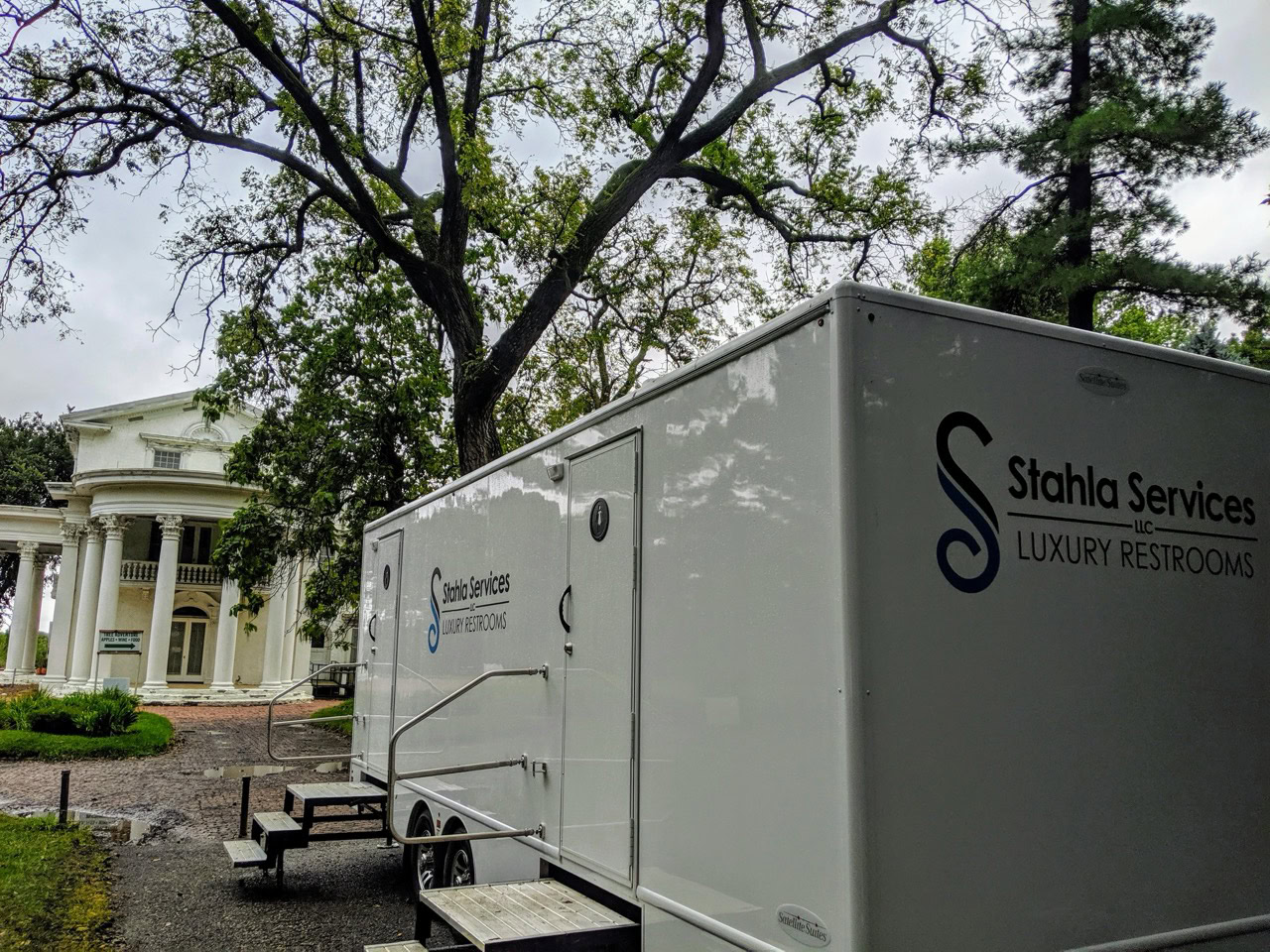 a 10 stall restroom trailer from stahla services is parked near a large, white, classical style building surrounded by trees.