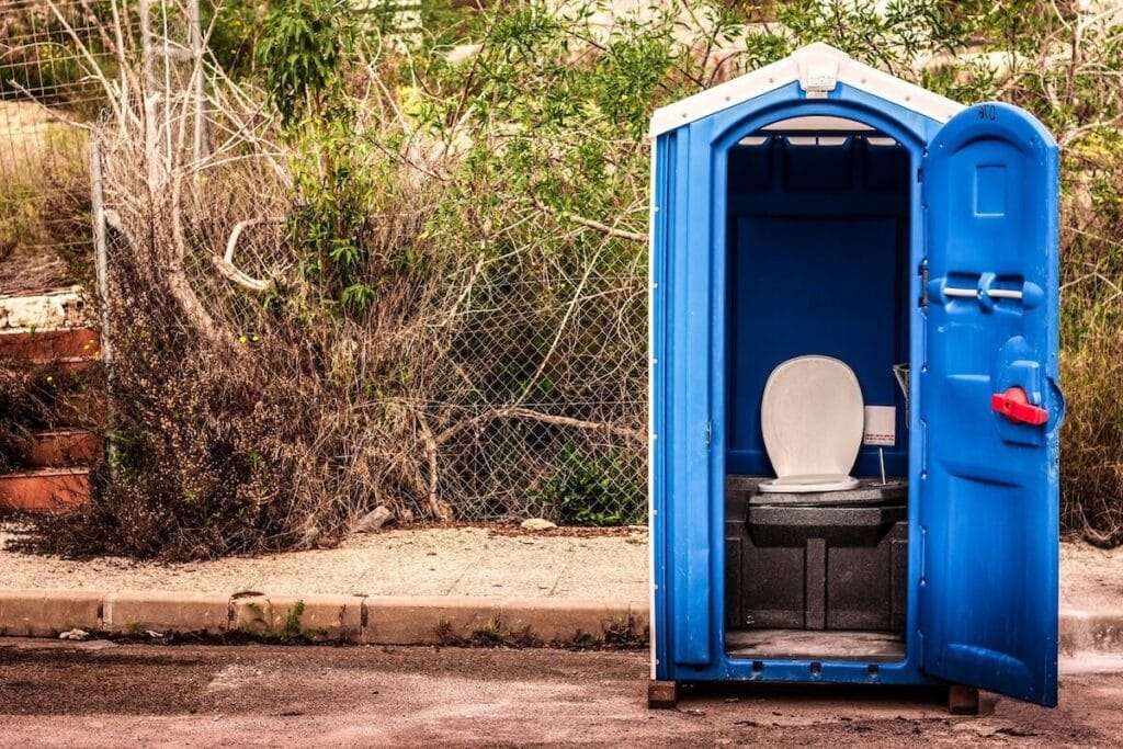 A flushable portable toilet with an open door, situated next to overgrown vegetation.
