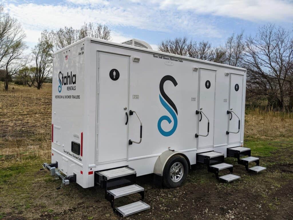 a white mobile restroom trailer with three separate stalls, labeled with the company name and logo "stahla rentals," is parked on a grassy area near a field, offering an essential 3 stall shower facility for outdoor events.