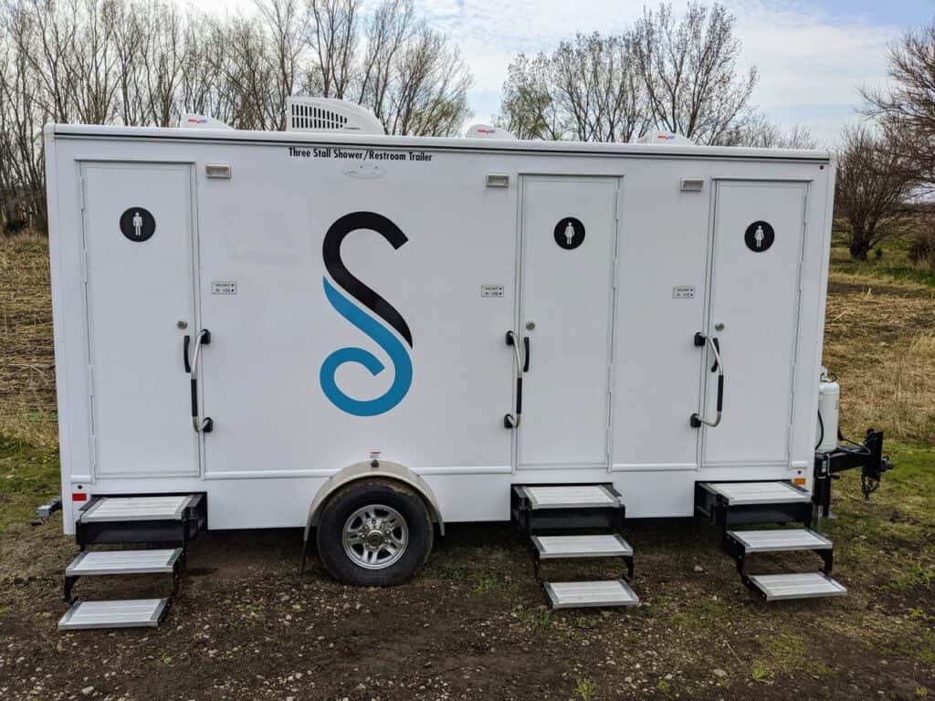 a white restroom trailer featuring a 3 stall shower setup with black and blue signage is parked on a grassy area, framed by trees. each stall is equipped with a step and has a clearly labelled door.